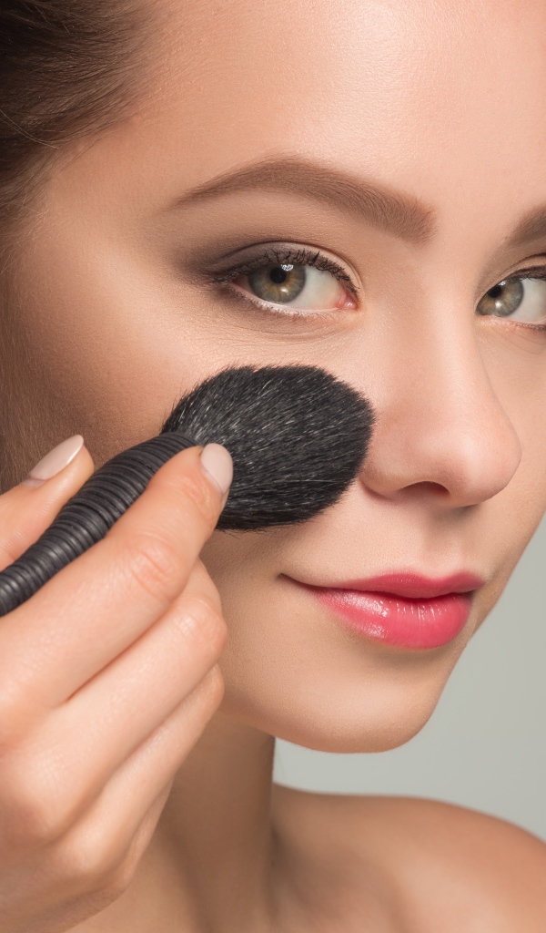 Красивой девушке наносят макияж на лицо кистью 