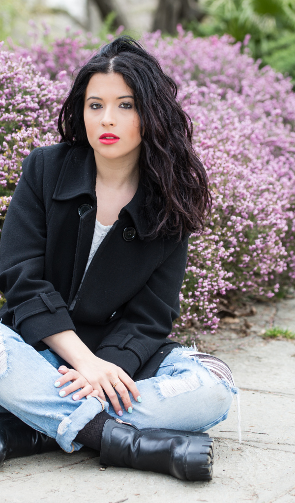 Красивая девушка в черном пальто сидит на земле у кустов