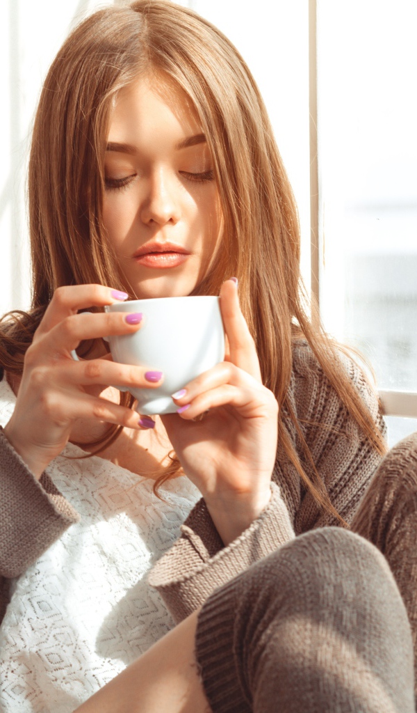 Красивая девушка в свитере пьет кофе у окна