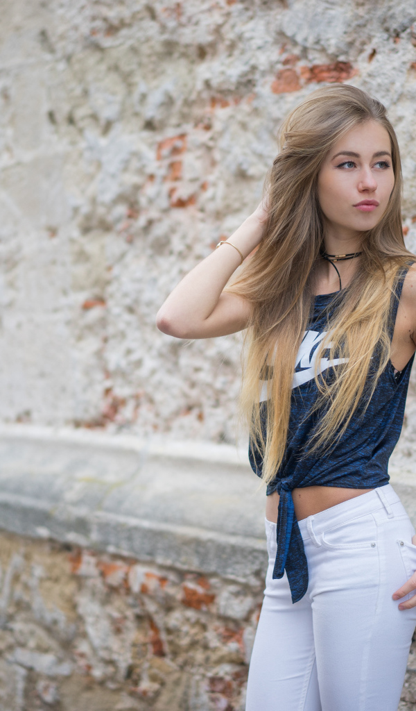 Красивая девушка в белых брюках стоит у стены 