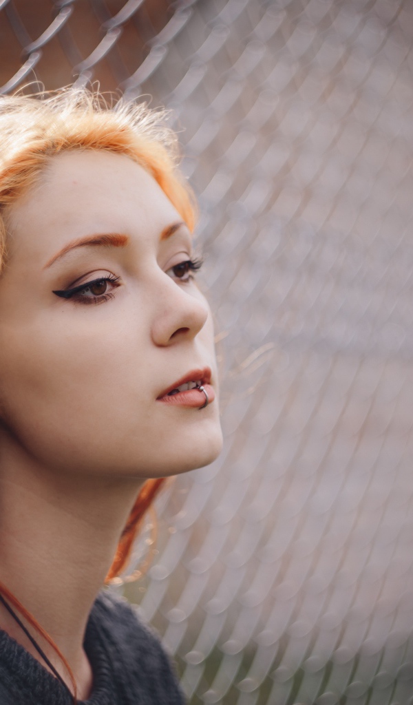 Красивая рыжеволосая девушка с пирсингом у железной сетки 