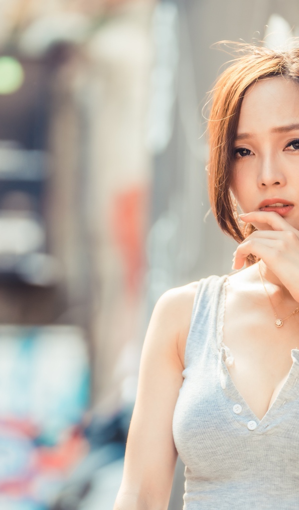 Милая девушка азиатка  с рукой у лица стоит у стены 