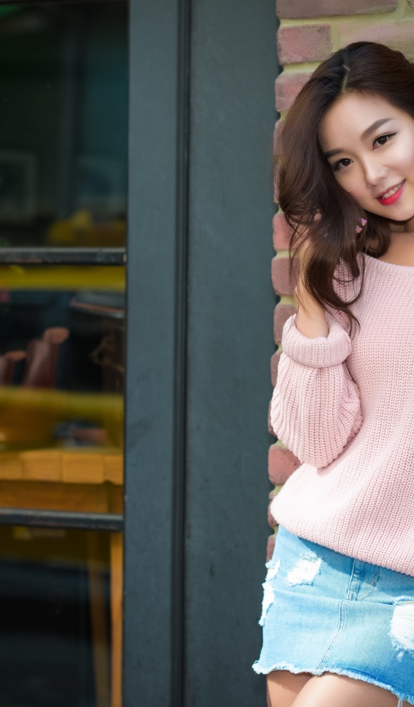 Милая азиатка в розовом свитере стоит у стены 