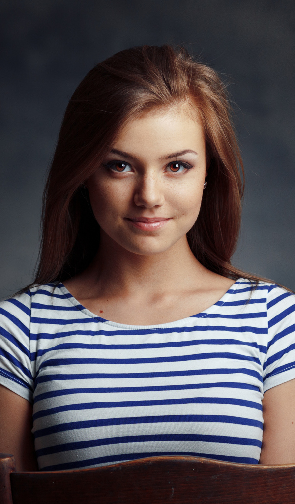 Милая кареглазая девушка в полосатой майке сидит на стуле 