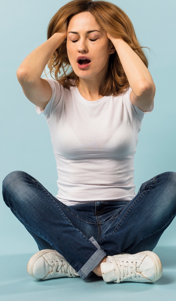 Девушка с руками в волосах сидит в джинсах на голубом фоне