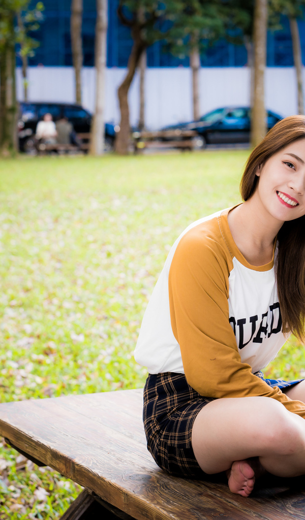 Улыбающаяся девушка азиатка сидит на лавке в парке 