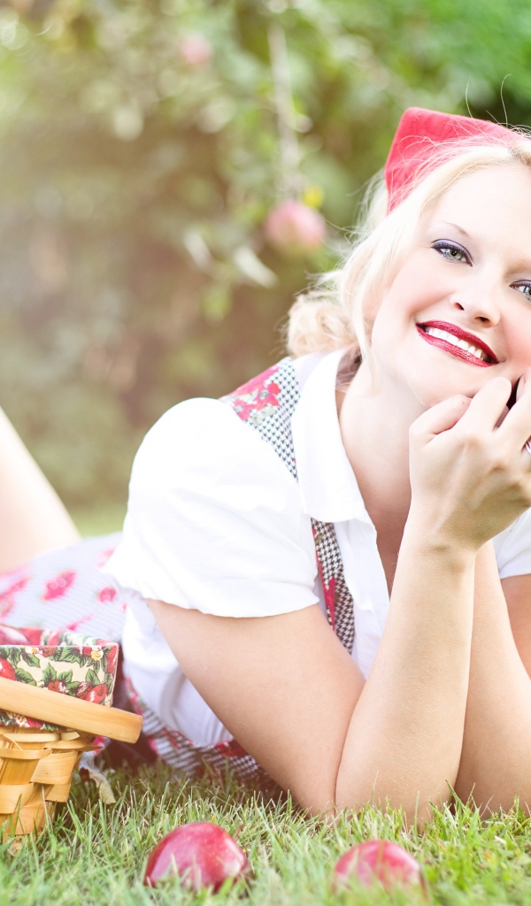Улыбающаяся блондинка лежит на зеленой траве с корзиной яблок 