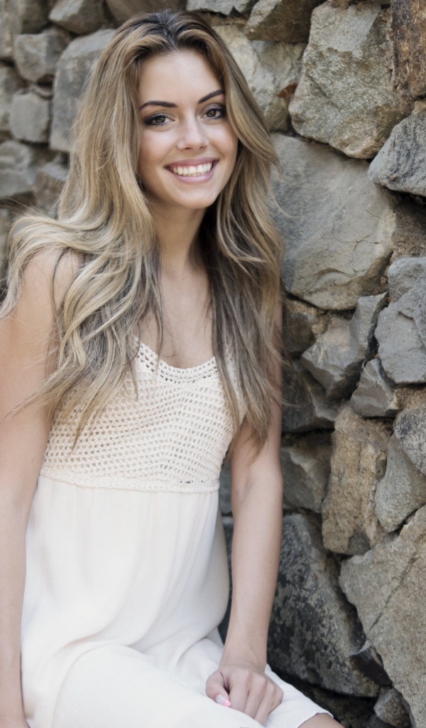 Улыбающаяся  девушка в белом платье стоит на ступеньках у стены