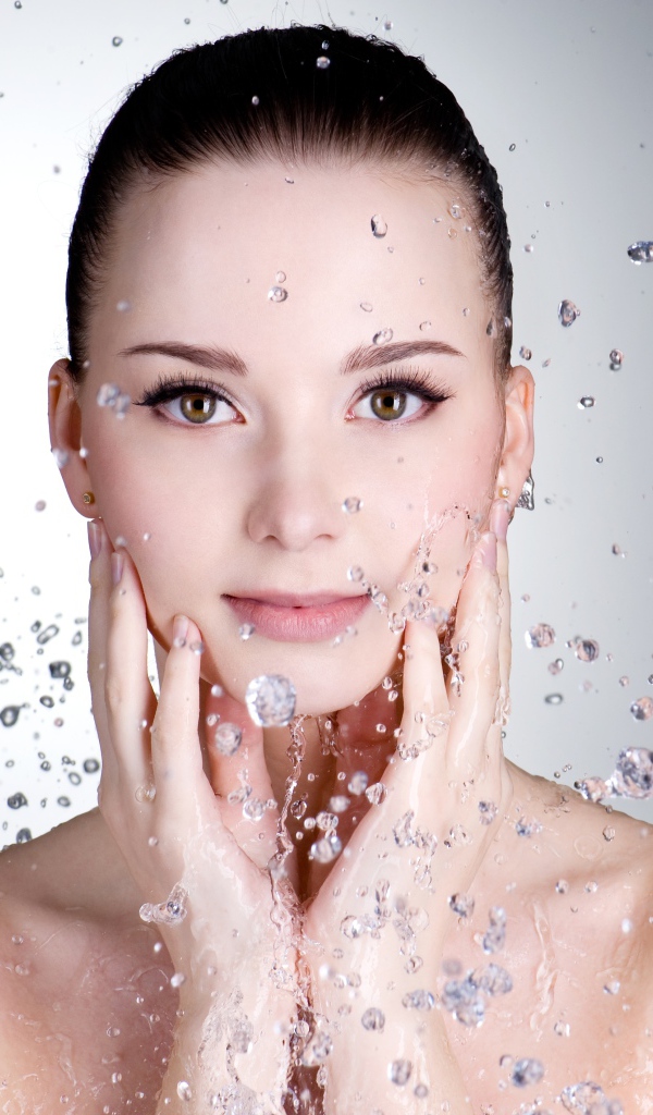 Брызги воды летят на красивое лицо молодой девушки на сером фоне