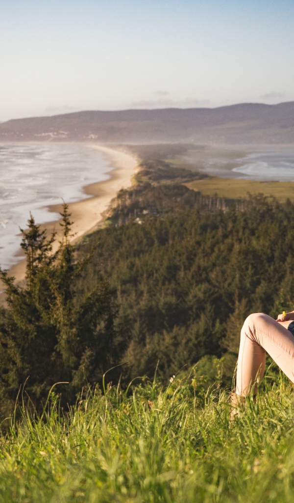 Молодая девушка сидит на зеленой траве на фоне моря