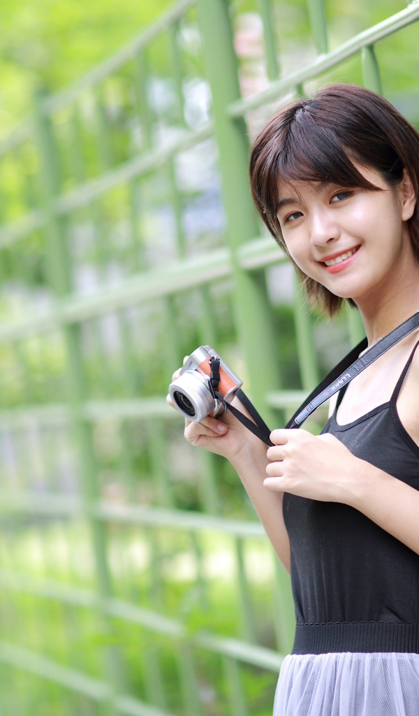 Молодая улыбающаяся азиатка с фотоаппаратом у забора 