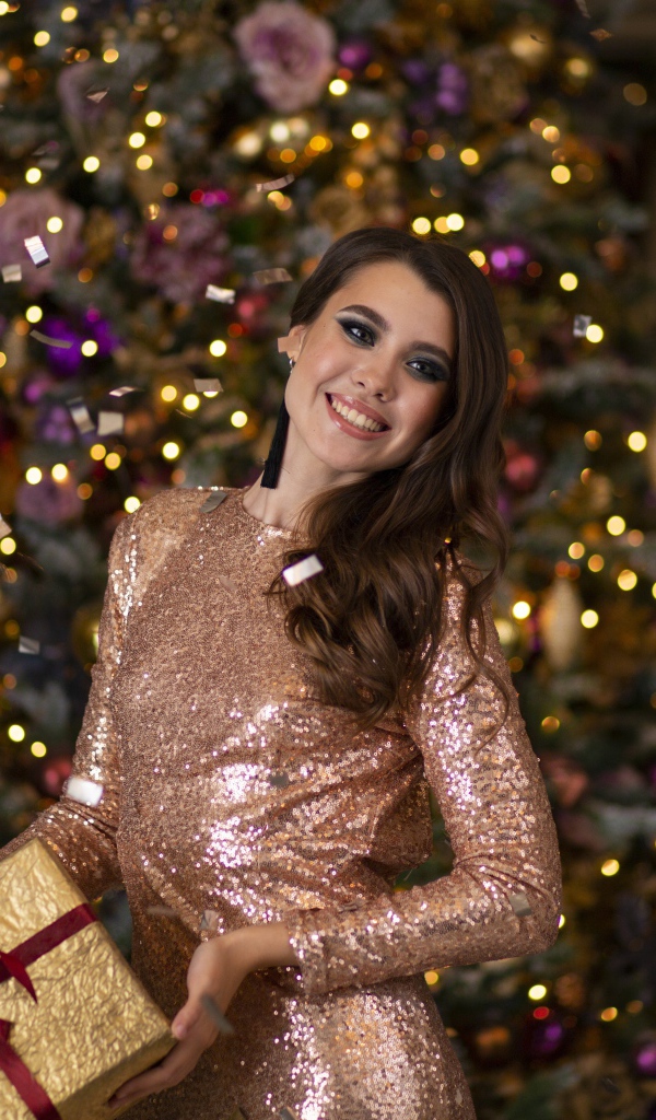 Милая улыбающаяся девушка в красивом платье у елки с подарком