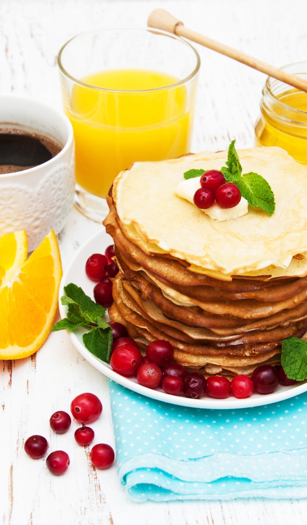 Тонкие румяные блинчики на столе с ягодами клюквы, апельсинами, медом и кофе