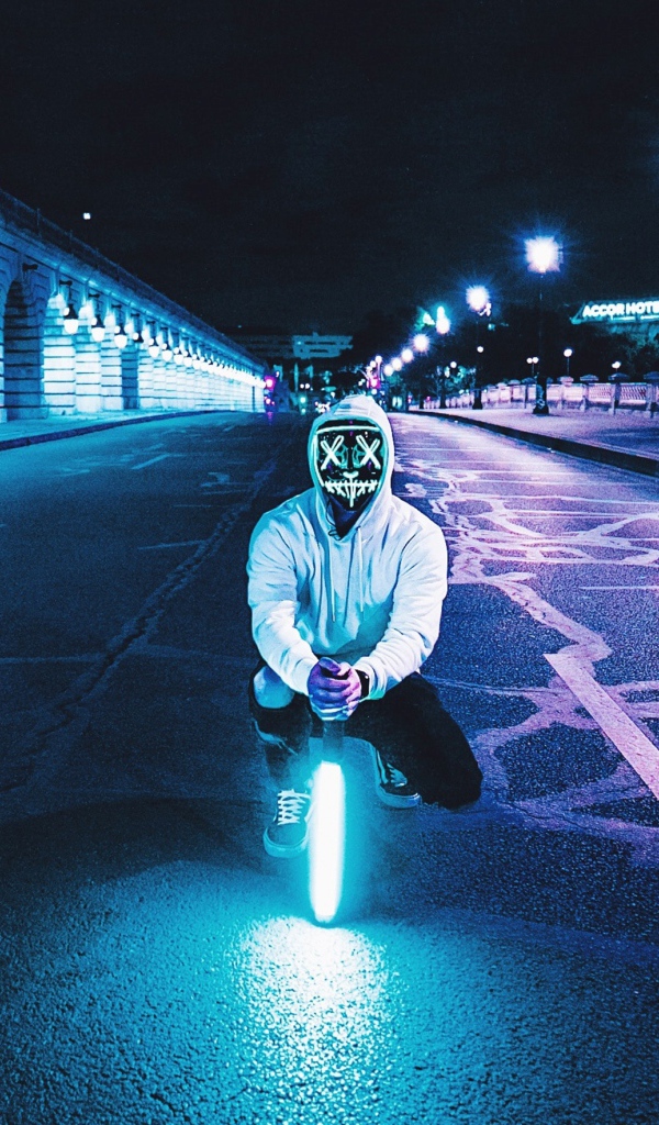 Парень в светящейся маске сидит с неоновой лампой на дороге