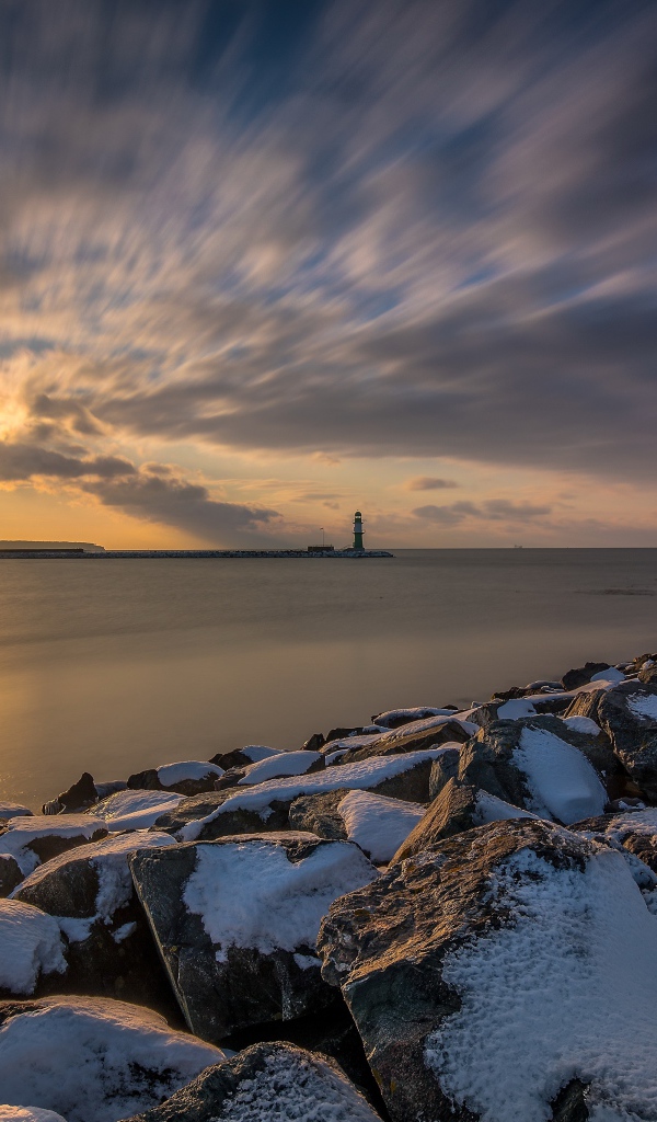Заснеженные камни на берегу моря с маяком на закате