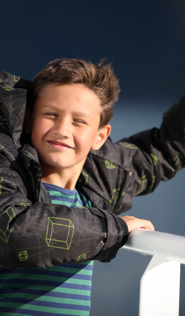 Веселый мальчик в куртке стоит на ветру в лучах солнца