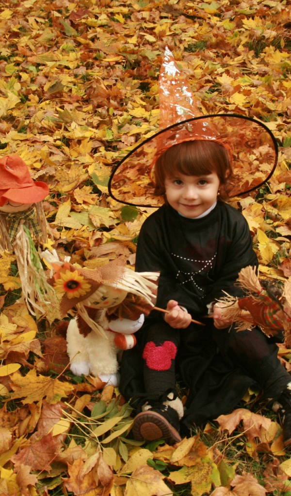 Маленькая девочка в костюме ведьмы сидит на опавшей листве на Хэллоуин