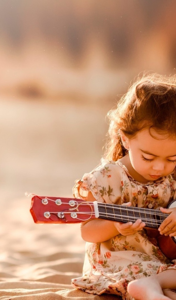 Маленькая девочка с гитарой сидит на песке 