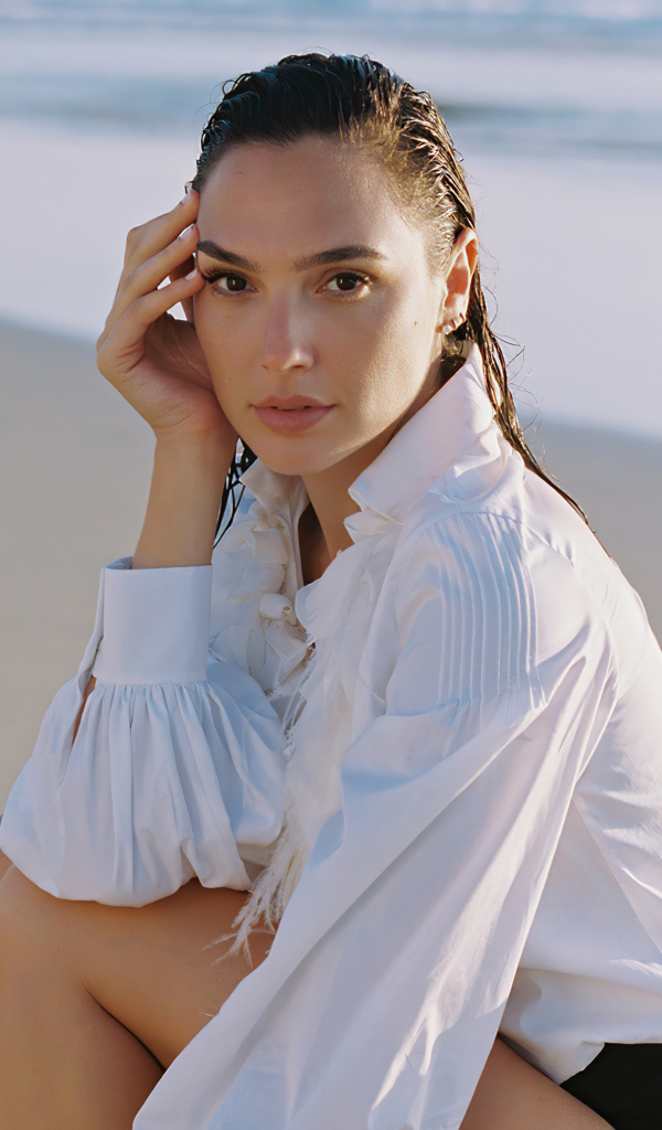 Актриса Галь Гадот в белой рубашке сидит на песке