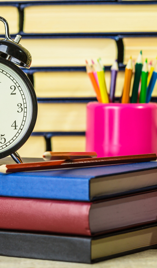 Будильник, книги, карандаши и яблоко на столе у стены 