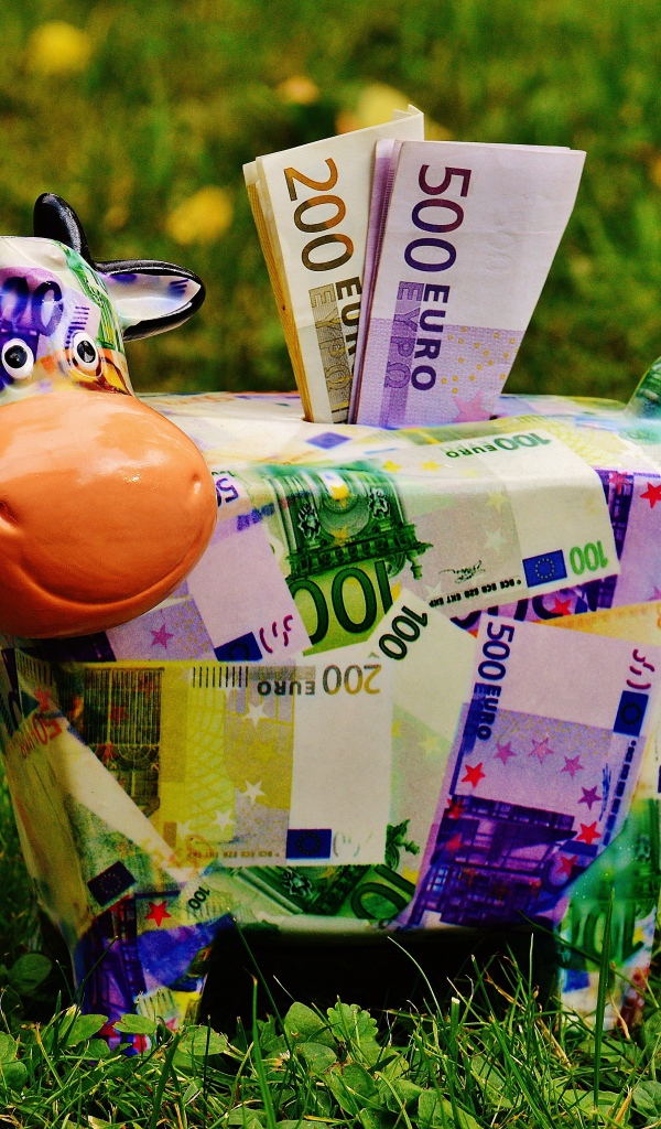 Копилка корова с купюрами евро на зеленой траве 