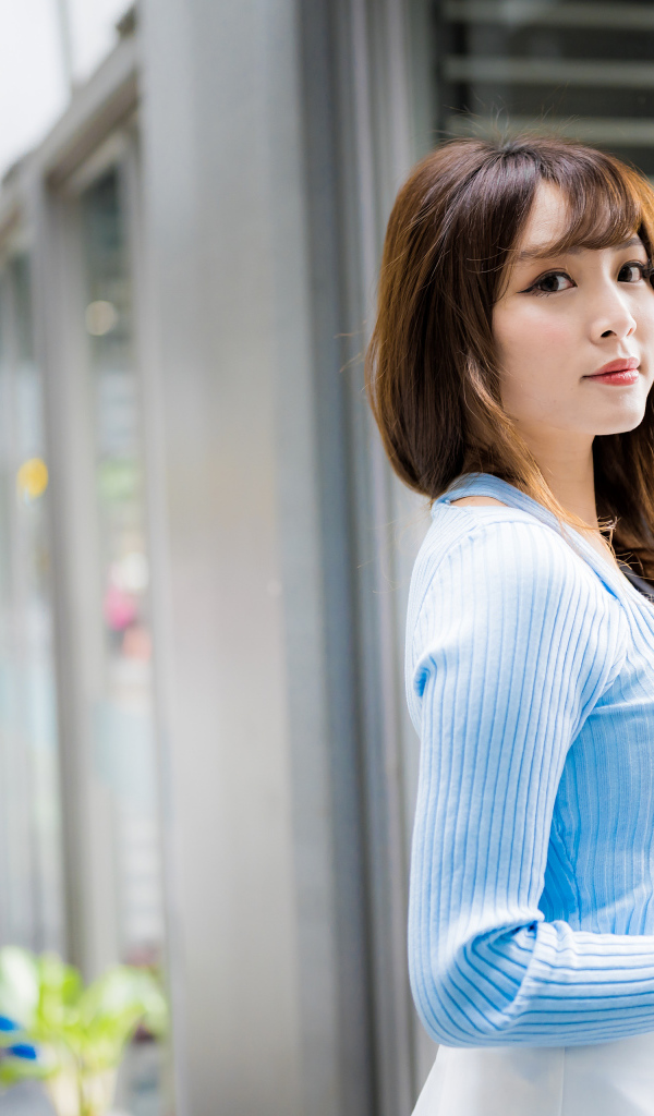 Красивая девушка азиатка в в голубой кофте стоит уокна