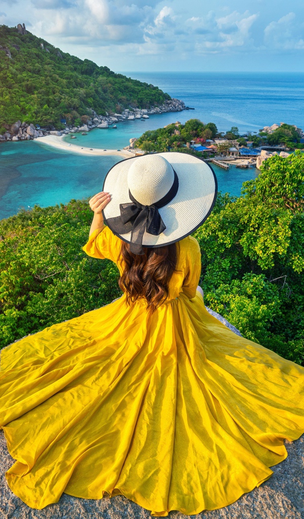 A girl in a beautiful yellow dress sits on a stone overlooking the sea