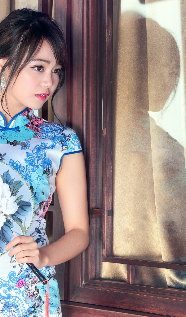 Девушка азиатка в красивом платье стоит у окна 