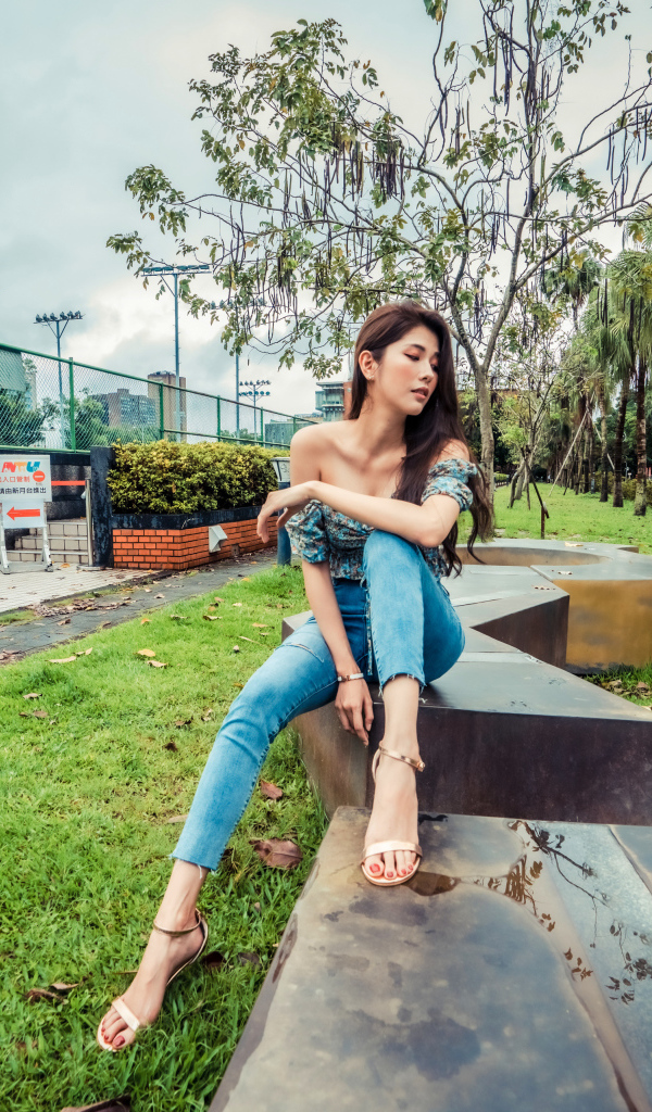 Девушка азиатка в джинсах сидит в парке 