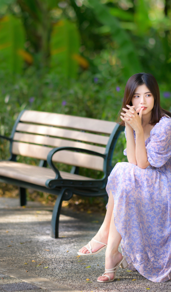 Грустная девушка азиатка в платье на лавке в парке 