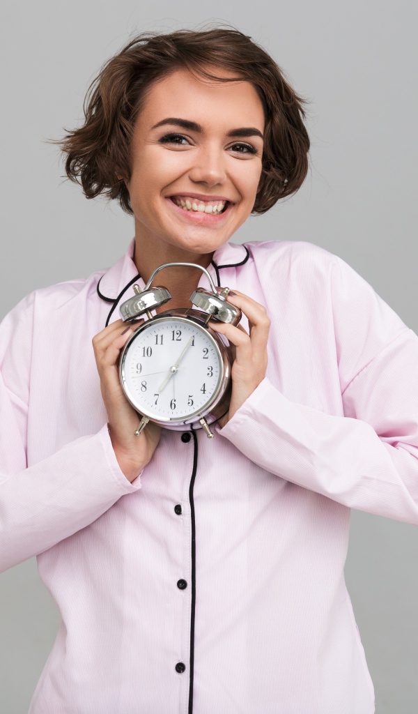 Улыбающаяся девушка в розовой рубашке с будильником в руках 