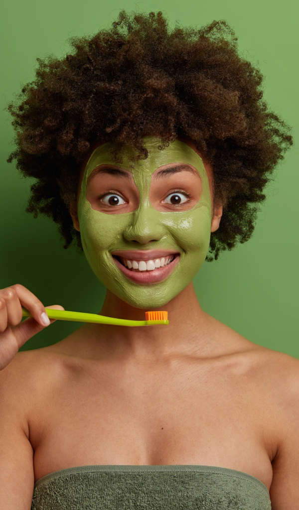 Улыбающаяся девушка с маской на лице чистит зубы