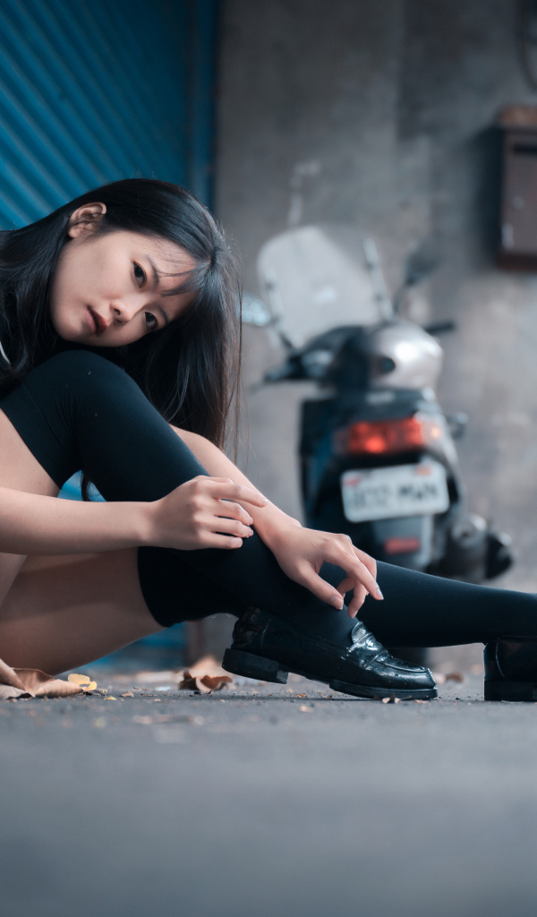 Молодая азиатка в черных чулках сидит у стены 