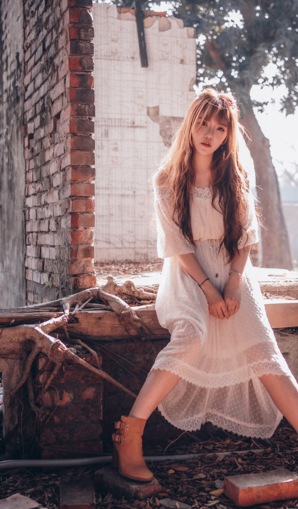 Молодая девушка азиатка в белом платье сидит на разваленной стене