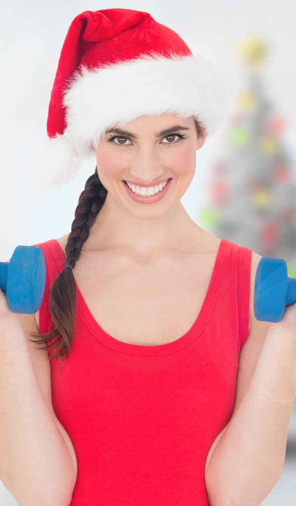 Красивая девушка в шапке Санта Клауса с гантелями в руках 