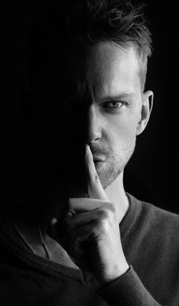 Молодой мужчина закрыл рот пальцем на черном фоне 