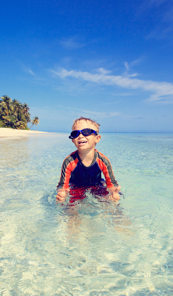 Мальчик в очках сидит в воде на пляже 