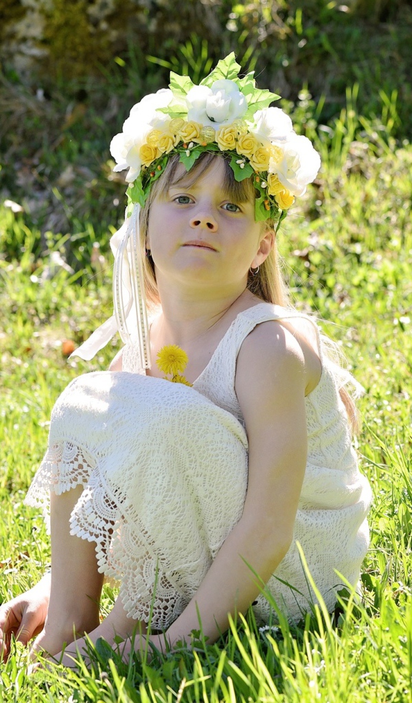 Маленькая девочка с венком на голове сидит на зеленой траве