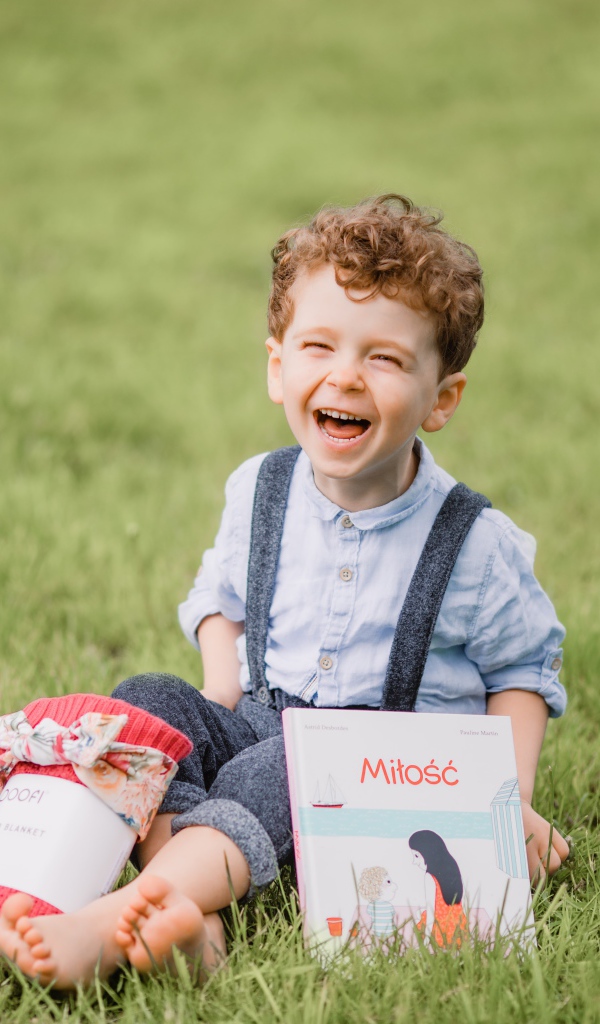Маленький улыбающийся мальчик сидит на зеленой траве с книгой 
