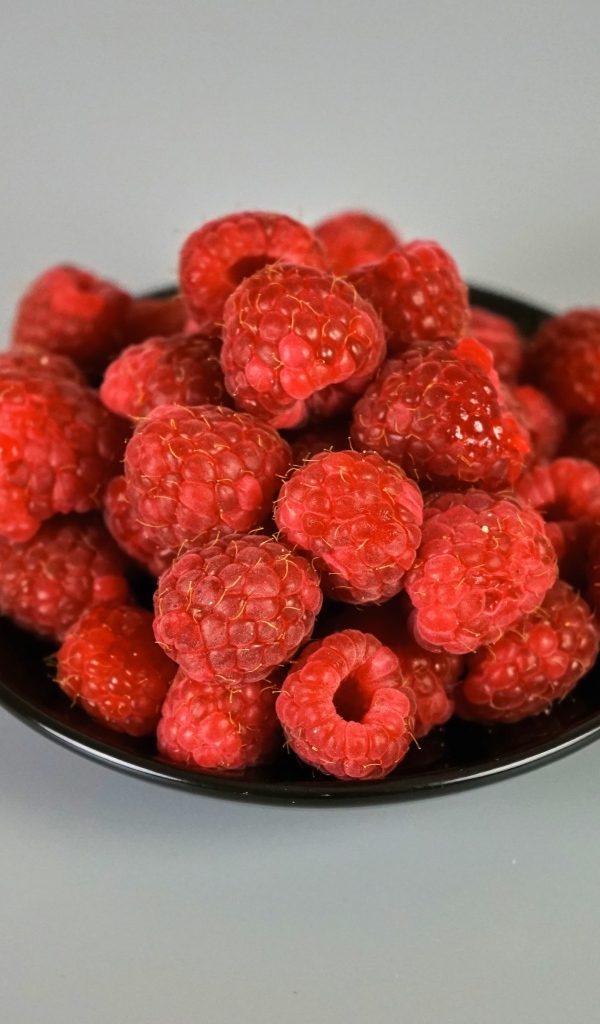 Спелая красная малина в черной тарелке на сером фоне