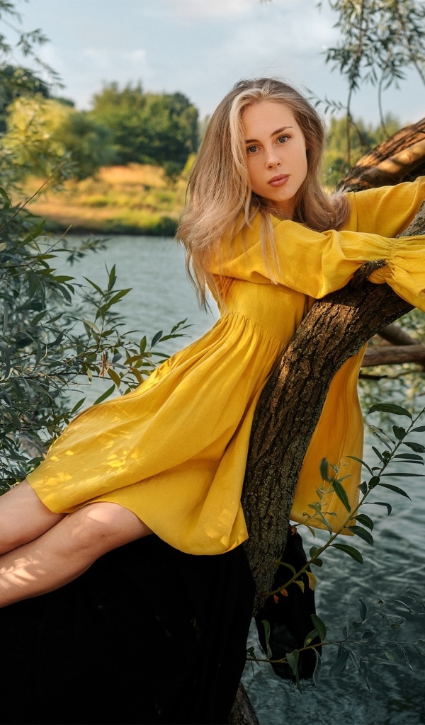Красивая девушка в желтом платье лежит на дереве у воды