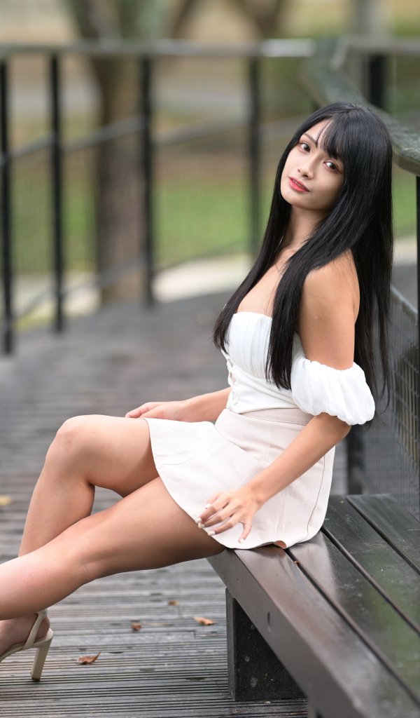 Красивая длинноволосая азиатка в белом платье сидит на лавке