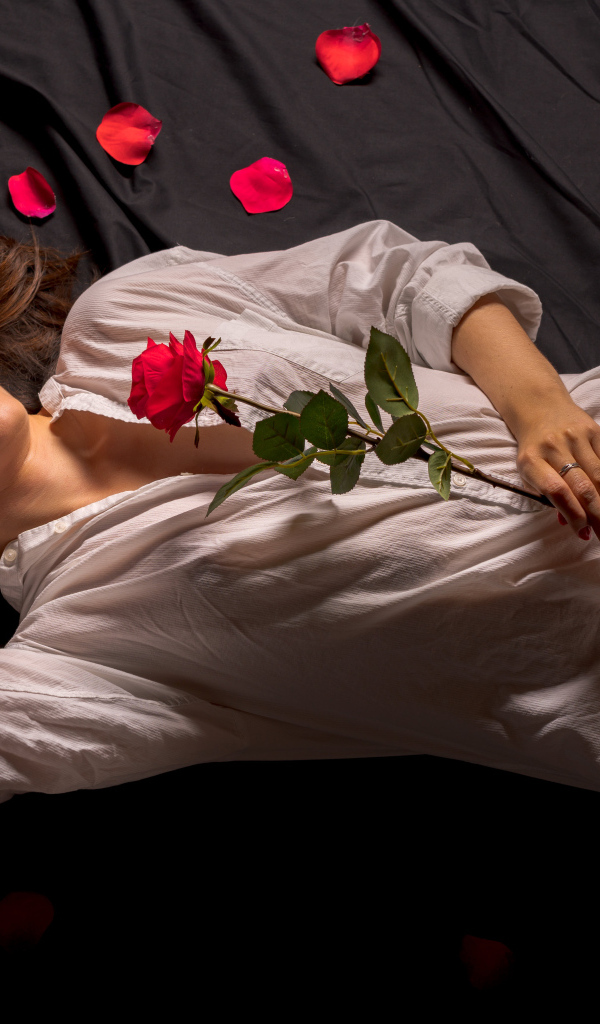 Девушка в белой рубашке с розой в руках лежит на постели