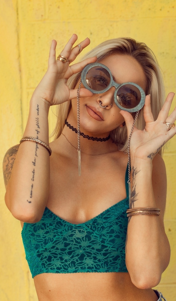 Девушка в очках с татуировками на руках стоит у стены