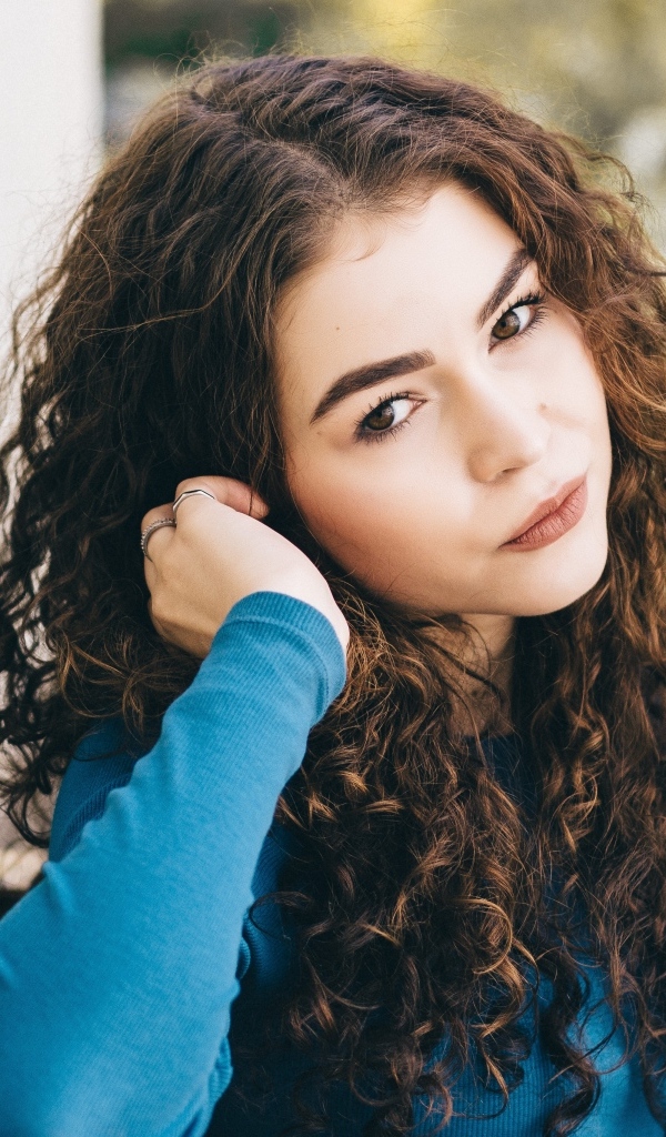 Кареглазая девушка с кудрявыми волосами стоит у стены