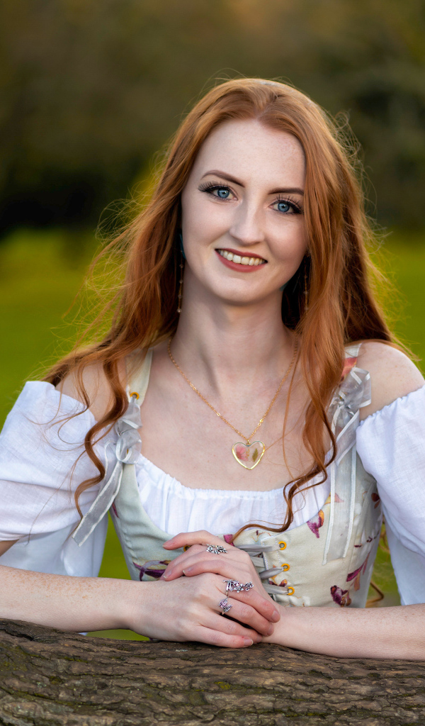 Улыбающаяся рыжеволосая девушка стоит у дерева