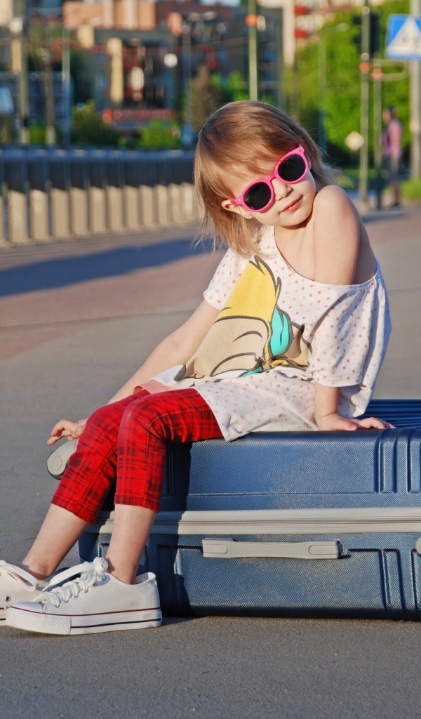Девочка в солнечных очках сидит на чемодане на дороге