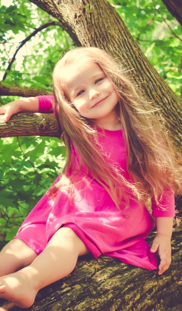 Милая девочка в розовом платье сидит на ветке дерева