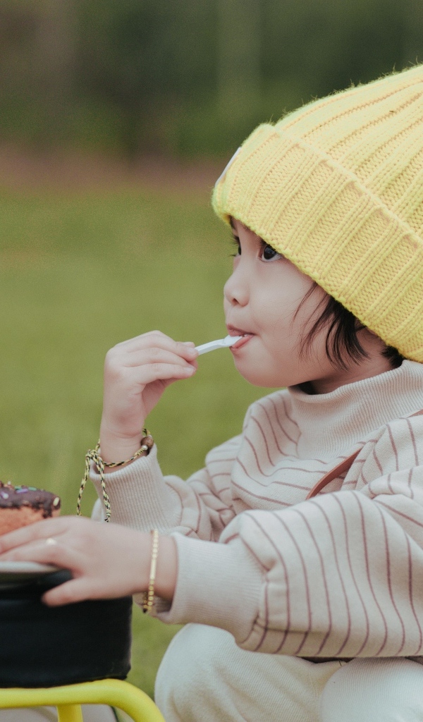 Маленькая девочка в желтой шапке ест пирожное