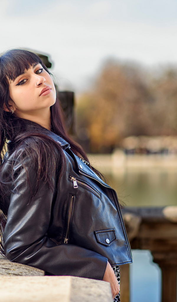 Девушка в черной куртке стоит на мосту у воды